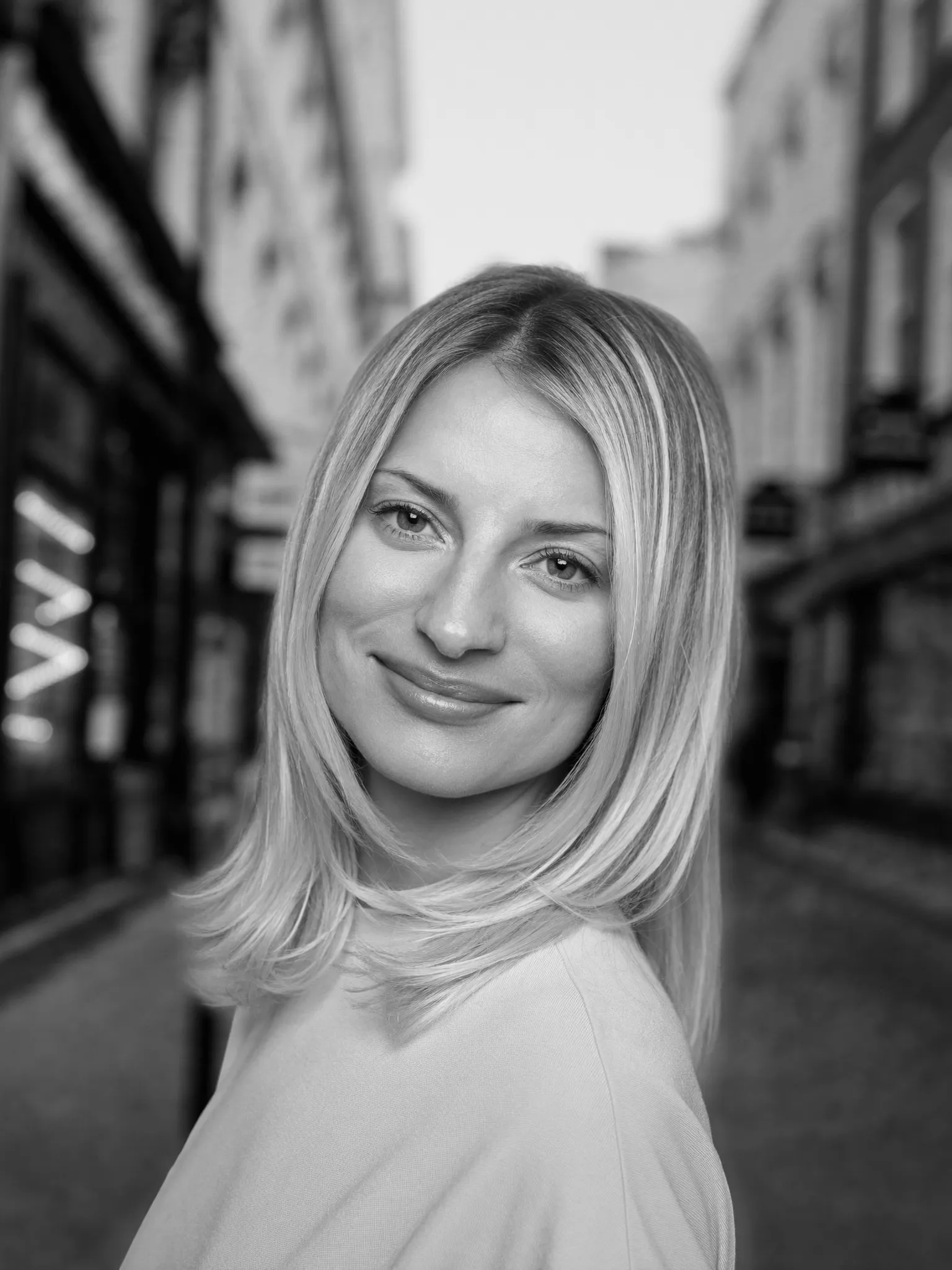 Lange laagjes — Zwart-witportret van een vrouw met lang haar in losse laagjes, driekwart naar de camera gedraaid. Onscherpe achtergrond van een Nederlandse straat.