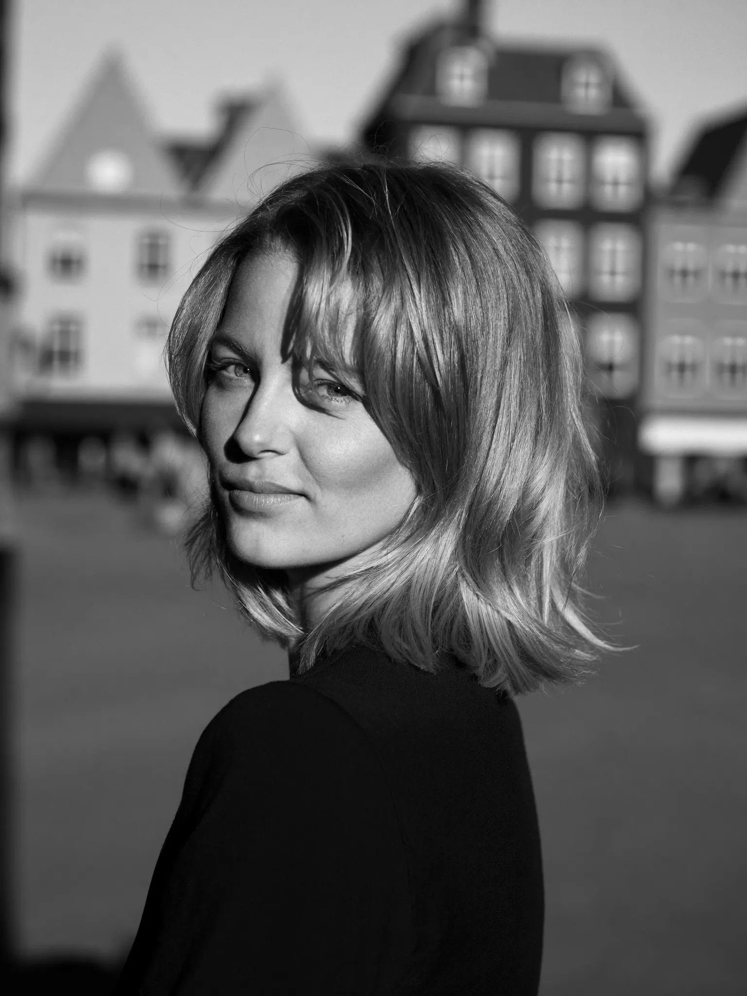Wolfcut — Zwart-witportret van een vrouw met een wolfcut: schoudherlang haar met sterke laagjes en een lichte pony. Onscherpe achtergrond van een Nederlands plein.